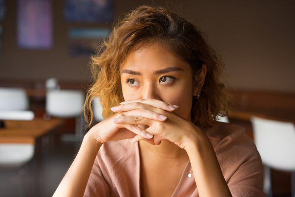 Closeup of worried young Asian woman at cafe covering nose and mouth with clasped hands.